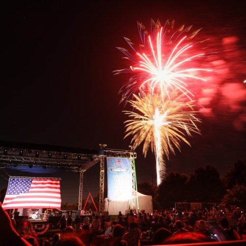 Fireworks illuminate the night sky above a stage with an American flag, celebrating a festive event.