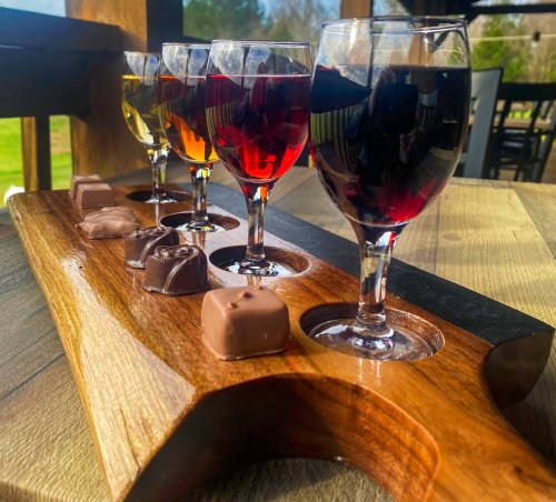 A wooden flight board with four glasses of colorful drinks and chocolate pieces, set against a scenic outdoor background.