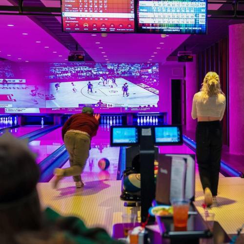 A bowling alley with a large screen showing a basketball game, featuring two bowlers and people seated at a table.