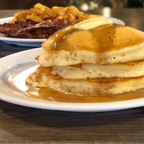 A stack of fluffy pancakes topped with butter and syrup, served with crispy bacon and golden hash browns.