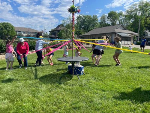 A group of children and adults participate in a maypole dance on a sunny day, surrounded by green grass and buildings.