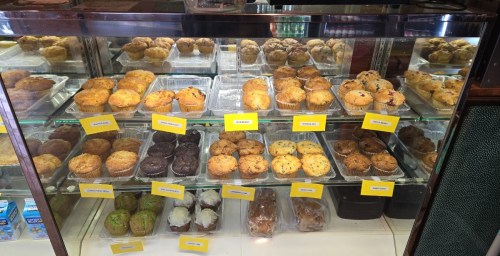 A display case filled with various muffins, labeled with names, showcasing a variety of flavors and colors.