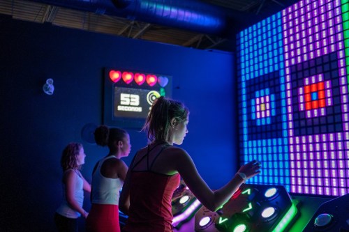 Three young people interact with a colorful, illuminated game display in a dark arcade setting.