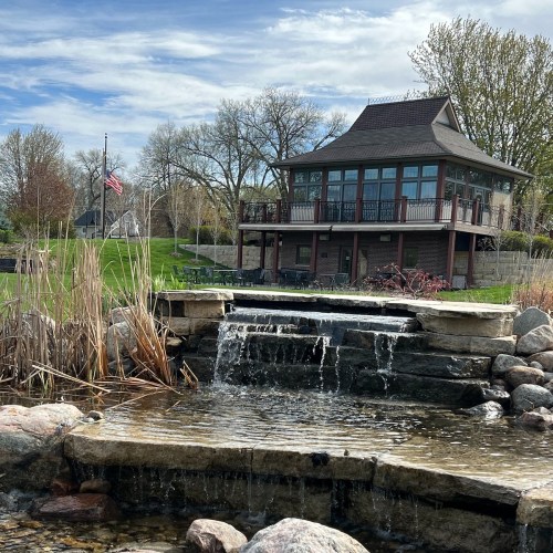 A serene scene featuring a waterfall in front of a modern house surrounded by trees and a clear blue sky.