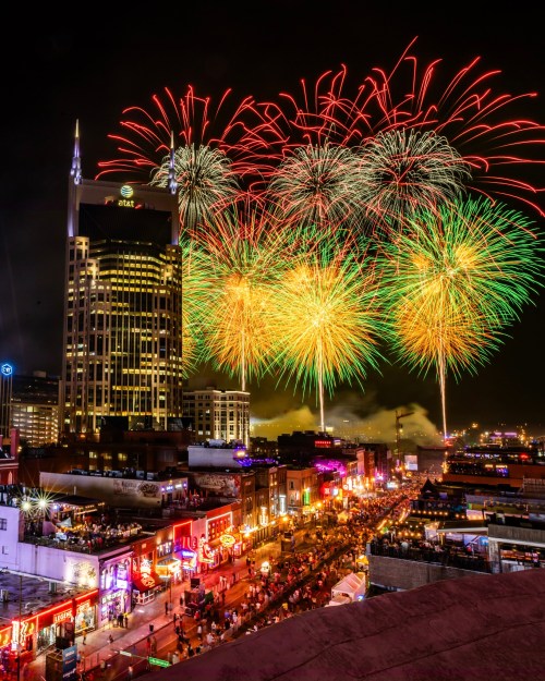Colorful fireworks illuminate the night sky over a bustling city street, with a tall building in the background.