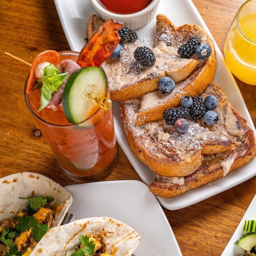 A plate of French toast topped with berries and powdered sugar, alongside a cocktail and tacos on a wooden table.