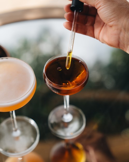 A hand holds a dropper above a cocktail glass, with two other drinks in the background on a reflective surface.