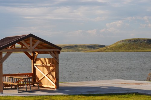 A wooden gazebo by a calm lake, with grassy hills in the background under a partly cloudy sky.