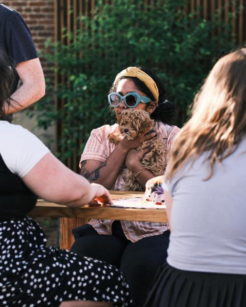 A woman wearing sunglasses and a headband holds a small dog while sitting at a table with others outdoors.