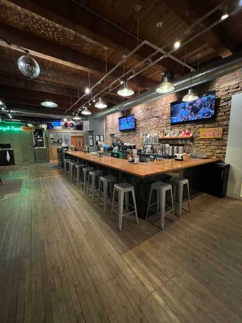 A spacious bar area with wooden floors, stone walls, and high stools, featuring TVs and a disco ball overhead.