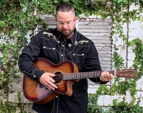 A man in a black western shirt plays an acoustic guitar against a backdrop of greenery and weathered wood.