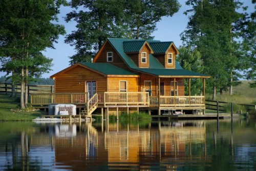 A charming wooden cabin with a green roof, surrounded by trees and reflecting in a calm lake.