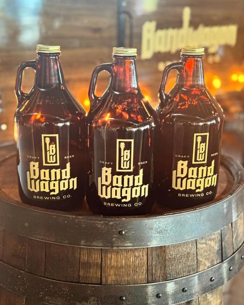 Three brown glass growlers labeled "Band Wagon Brewing Co." sit on a wooden barrel against a rustic backdrop.