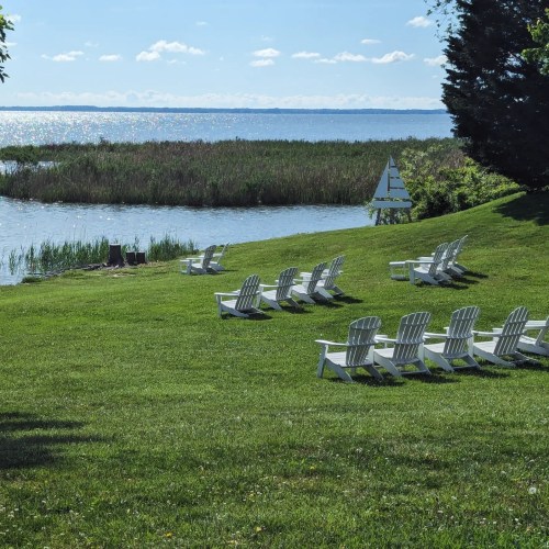 A serene lakeside scene with green grass, white Adirondack chairs, and a small sailboat in the distance under a clear sky.