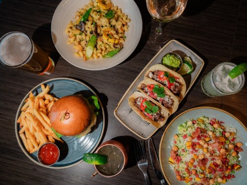 A table spread featuring pasta, tacos, a burger with fries, a salad, and drinks in a cozy setting.