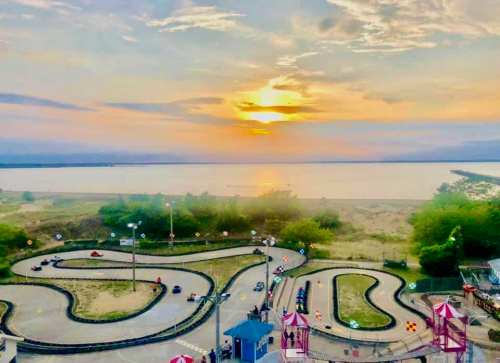 A scenic view of a go-kart track winding along a lake at sunset, with colorful structures in the foreground.