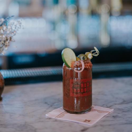 A vibrant cocktail in a glass jar, garnished with lime, olives, and herbs, sits on a marble bar top.