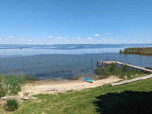 A serene lakeside view with a wooden dock, sandy shore, and a kayak on the grass under a clear blue sky.