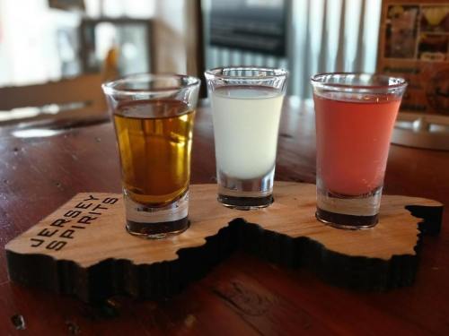 Three shot glasses on a wooden board shaped like New Jersey, featuring a golden drink, a clear drink, and a pink drink.