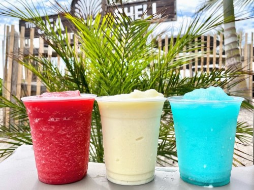Three colorful frozen drinks in clear cups: red, white, and blue, with a palm tree in the background.