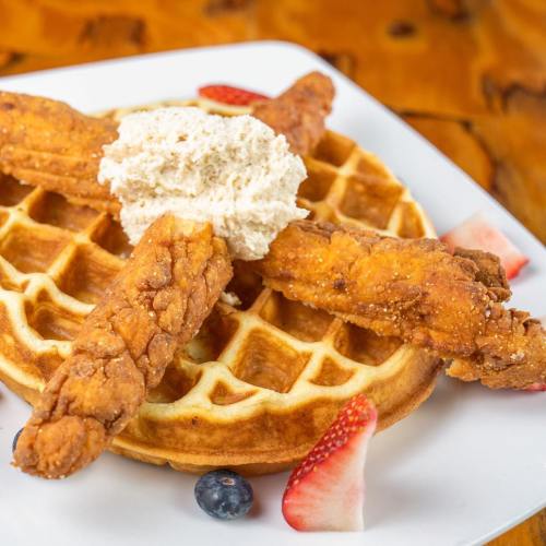 A plate featuring a golden waffle topped with fried chicken strips and a dollop of whipped cream, garnished with berries.