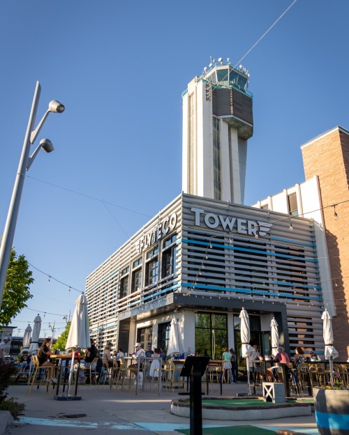 A modern building with "FlyteCo Tower" signage, outdoor seating, and a tall tower in the background under a clear blue sky.