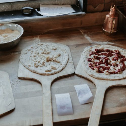 Two wooden pizza peels with dough, one plain and one topped with sauce and cheese, on a countertop.