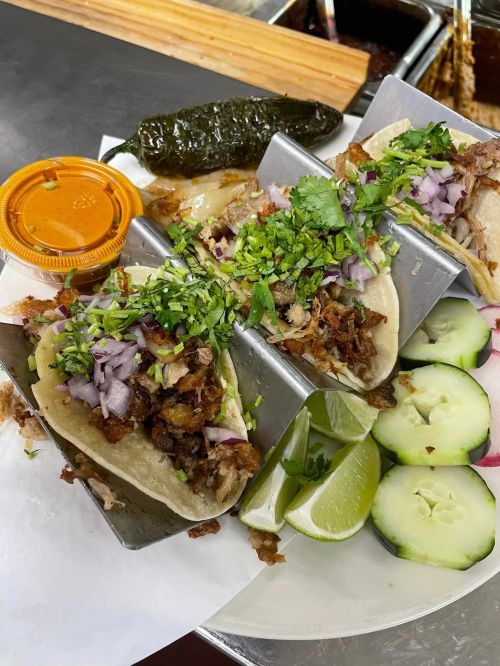 A plate of tacos topped with cilantro and onions, served with lime, cucumber slices, and a side of salsa and jalapeño.