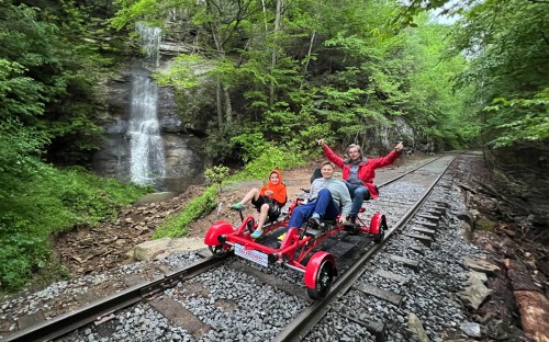 Three people enjoy a red rail bike on a scenic railway track near a waterfall and lush green trees.