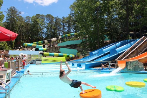 A water park scene with a child on a floating mat, colorful slides, and people enjoying the pool on a sunny day.