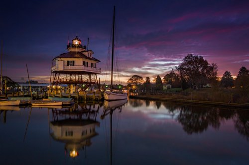 A lighthouse on stilts at dusk, reflecting in calm water, with boats docked nearby and a colorful sky.