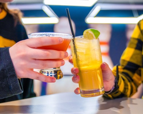 Two hands clink colorful cocktails together, one in a glass with a lime slice and the other with a salt rim.