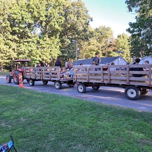 A tractor pulls two wooden wagons filled with people along a grassy path in a park setting.