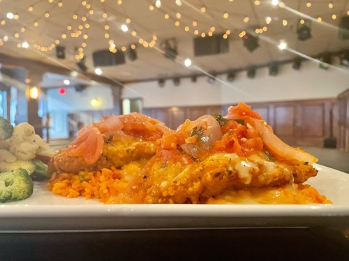 A plate of chicken topped with tomato sauce and onions, served with rice and mixed vegetables in a softly lit venue.