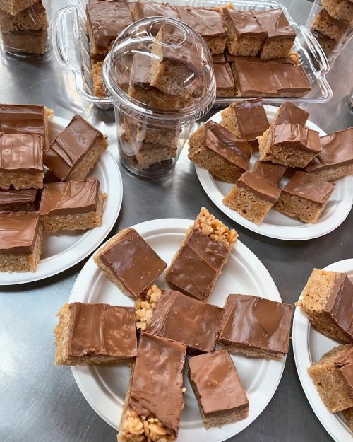 Plates of chocolate-covered rice crispy treats, with some in a clear plastic container on a silver surface.