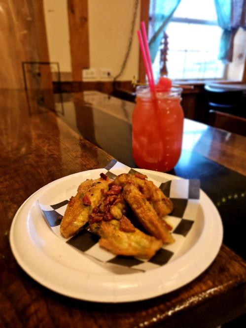 A plate of crispy fried pickles topped with bacon, next to a colorful drink with two straws on a wooden table.