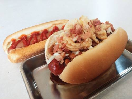 Two hot dogs on a tray: one topped with beans, bacon bits, and crispy onions; the other with ketchup.