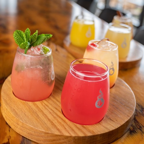 A wooden platter holds colorful cocktails in glass tumblers, garnished with mint and fruit, set on a table.
