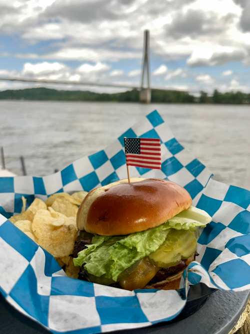 A burger with lettuce, pickles, and an American flag on top, served with chips, against a river backdrop.