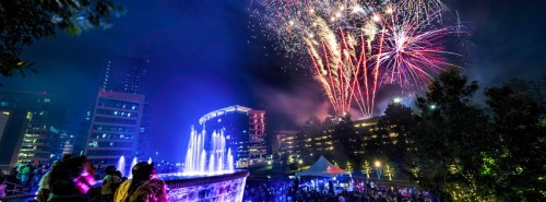 Colorful fireworks illuminate the night sky above a fountain and crowd at a festive outdoor event.