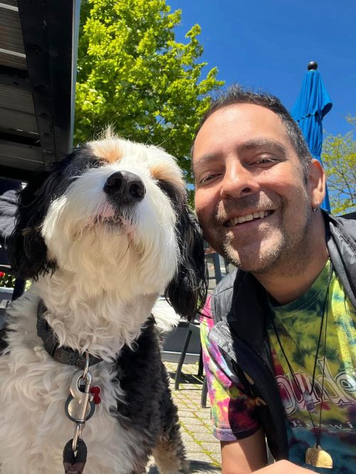 A smiling person poses with a fluffy dog outdoors, surrounded by greenery and a clear blue sky.