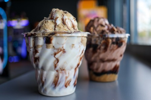 Two ice cream cups, one with chocolate and the other with vanilla swirls, topped with cookie crumbles and chocolate sauce.