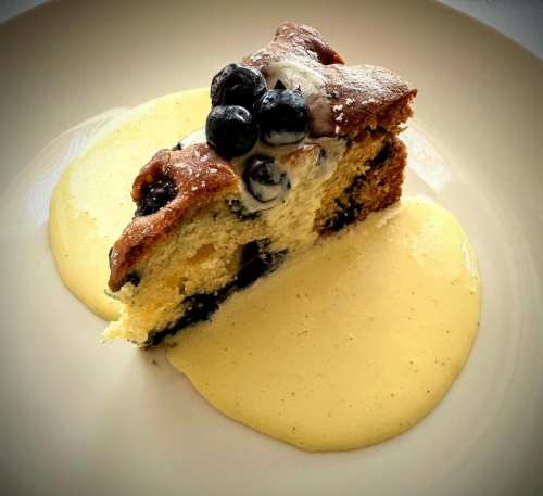 A slice of blueberry cake topped with cream, served on a plate with a drizzle of custard sauce.