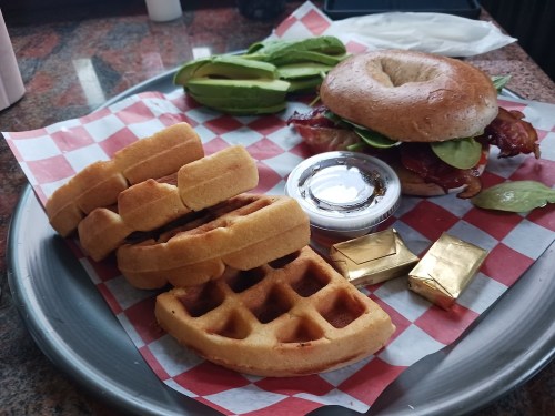 A tray with waffles, a bagel sandwich with bacon and greens, avocado slices, and a small container of syrup.