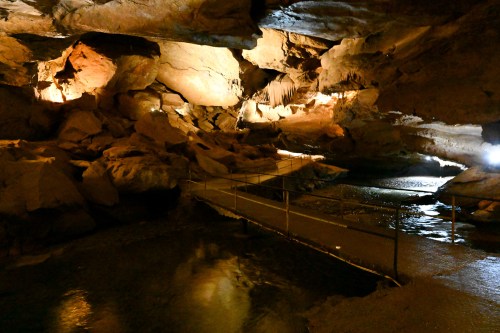 A dimly lit cave interior with rock formations, a water stream, and a walkway illuminated by soft lights.