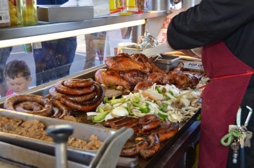 A vendor prepares various grilled meats and vegetables at a food stall, with a child peeking from the side.