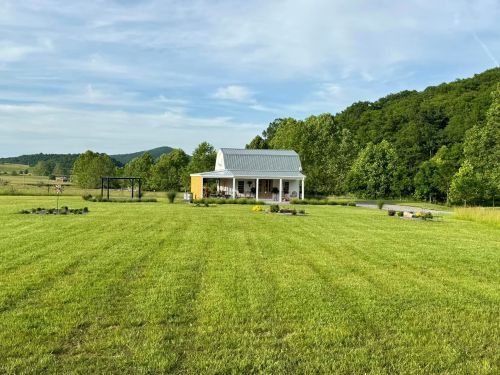 A small white house with a metal roof sits in a green field, surrounded by trees and rolling hills under a blue sky.
