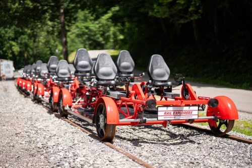 A row of red rail bikes with gray seats parked on a gravel track surrounded by greenery.