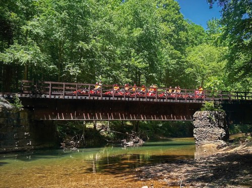 A wooden bridge spans a clear stream, surrounded by lush green trees, with people sitting on the bridge enjoying the view.