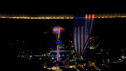 A night scene featuring fireworks and a large American flag draped from a bridge, illuminating the festive atmosphere below.